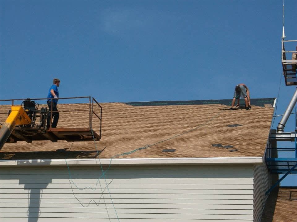 Two crew members on a boom lift, one on the platform and one on the roof, finishing a shingle ridge